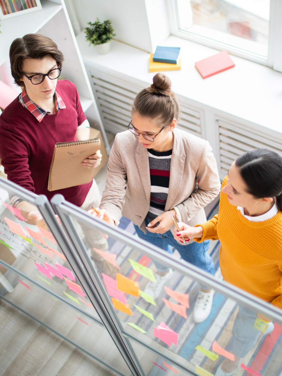 Group of three office workers standing around a glass board. Working on arranging sticky notes.
